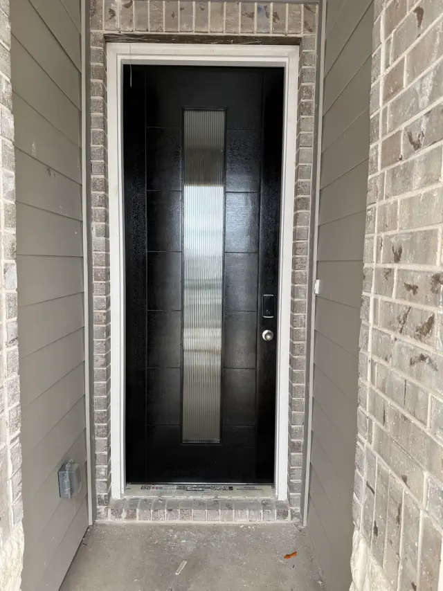 Modern black entry door with a tall narrow glass insert, installed in a brick entryway